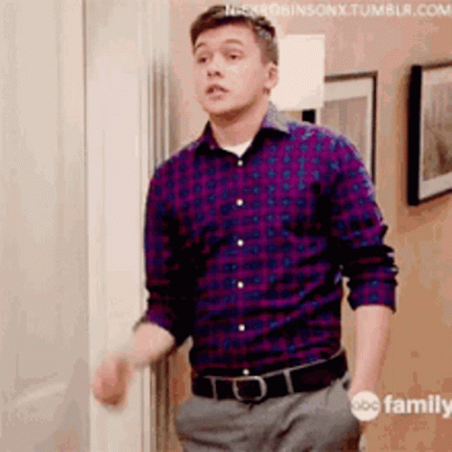 A Man In A Plaid Shirt And Gray Pants Is Standing In Front Of A Door . GIF
