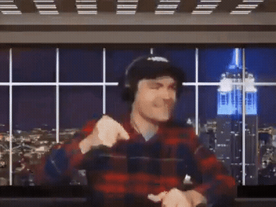A Man In A Plaid Shirt And Headphones Is Raising His Arms In The Air In Front Of A Large Screen . GIF