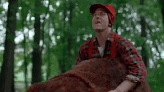 A Man In A Plaid Shirt And Red Hat Is Holding A Large Brown Bear In The Woods . GIF