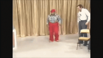 A Man In A Plaid Shirt And Red Pants Stands Next To A Clown GIF