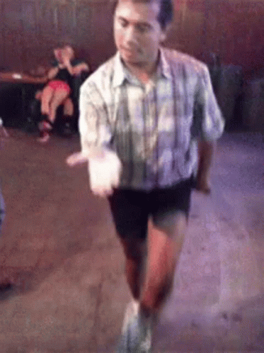 A Man In A Plaid Shirt And Shorts Is Dancing In A Room With People In The Background . GIF