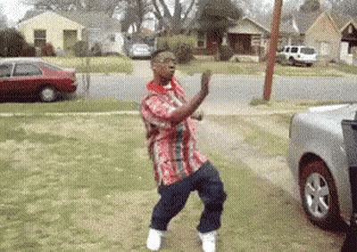 A Man In A Plaid Shirt Is Dancing In Front Of A Silver Truck . GIF