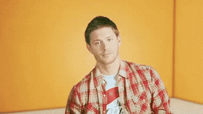 A Man In A Plaid Shirt Is Giving Two Thumbs Up In Front Of An Orange Wall . GIF