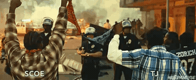 A Man In A Plaid Shirt Is Holding A Bandana In Front Of Riot Police . GIF