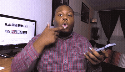 A Man In A Plaid Shirt Is Holding A Cell Phone In Front Of A Computer Monitor GIF