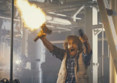 A Man In A Plaid Shirt Is Holding A Flamethrower In His Right Hand GIF