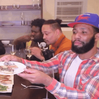 A Man In A Plaid Shirt Is Holding A Plate Of Food In Front Of A Group Of Men GIF