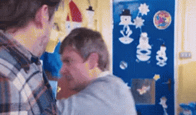 A Man In A Plaid Shirt Is Holding Another Man In His Arms In Front Of A Blue Wall With Snowmen On It . GIF
