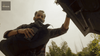 A Man In A Plaid Shirt Is Reaching Into The Trunk Of A Car With A Sign Above Him That Says Mumble GIF