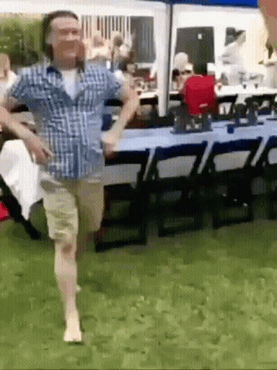 A Man In A Plaid Shirt Is Running Barefoot In The Grass In Front Of A Table And Chairs . GIF