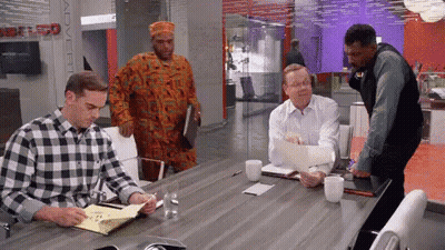 A Man In A Plaid Shirt Is Sitting At A Conference Table With Other Men GIF