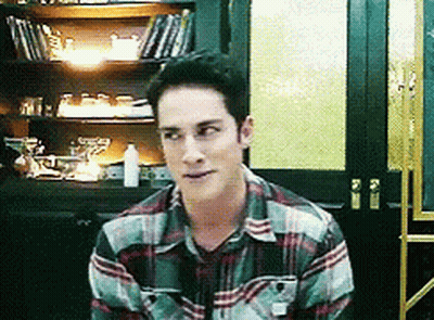 A Man In A Plaid Shirt Is Sitting In Front Of A Bookcase GIF