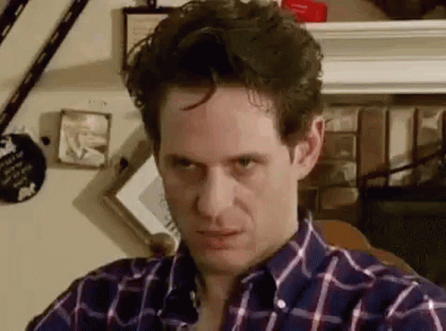A Man In A Plaid Shirt Is Sitting In Front Of A Fireplace And Looking At The Camera . GIF