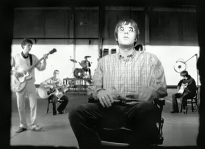 A Man In A Plaid Shirt Is Sitting In Front Of A Group Of Musicians GIF