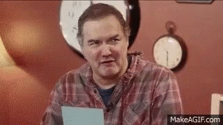 A Man In A Plaid Shirt Is Sitting In Front Of A Wall With Clocks . GIF