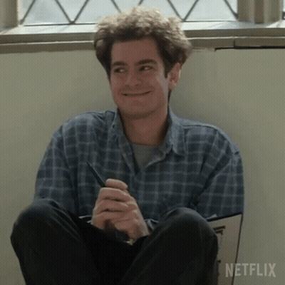 A Man In A Plaid Shirt Is Sitting On The Floor Holding A Pen And Smiling . GIF