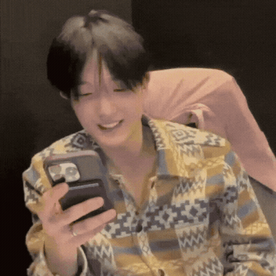 A Man In A Plaid Shirt Is Smiling While Holding A Cell Phone In His Hand . GIF