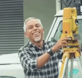 A Man In A Plaid Shirt Is Smiling While Holding A Yellow Device On A Tripod GIF