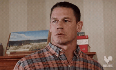 A Man In A Plaid Shirt Is Standing In Front Of A Stack Of Books And A Painting . GIF