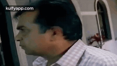 A Man In A Plaid Shirt Is Standing In Front Of A Window And Looking At Something . GIF