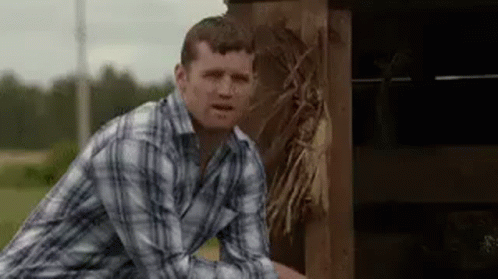 A Man In A Plaid Shirt Is Standing Next To A Wooden Fence And Making A Funny Face . GIF