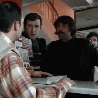 A Man In A Plaid Shirt Is Talking To Another Man In Front Of A Sign That Says T.i. GIF