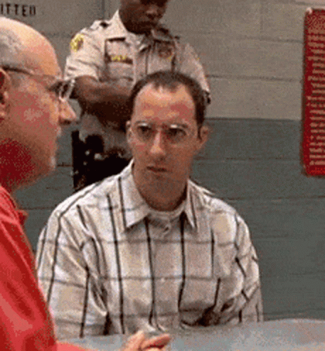 A Man In A Plaid Shirt Sits At A Table Talking To Another Man While A Police Officer Stands Behind Him GIF