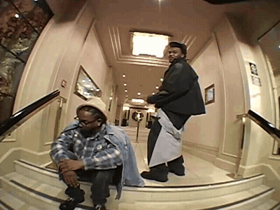 A Man In A Plaid Shirt Sits On A Set Of Stairs Next To Another Man In A Black Jacket GIF