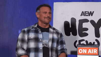 A Man In A Plaid Shirt Stands In Front Of A Sign That Says Aew Hey GIF