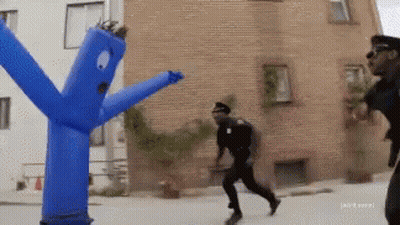 A Man In A Police Uniform Is Running Away From A Blue Blow Up Man GIF