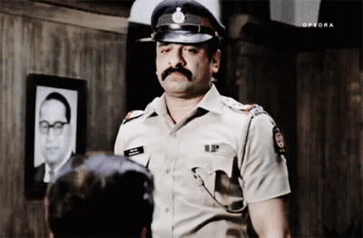 A Man In A Police Uniform Is Standing In Front Of A Picture Of A Man With A Mustache GIF