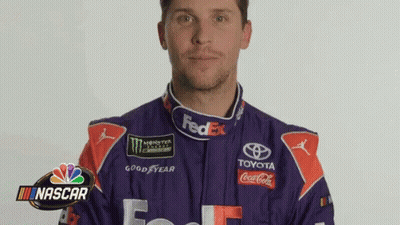 A Man In A Purple And Red Racing Suit Is Standing In Front Of A Nascar Sign . GIF