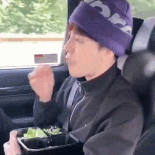 A Man In A Purple Hat Is Sitting In The Back Seat Of A Car Eating A Salad . GIF