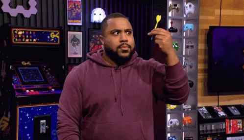 A Man In A Purple Hoodie Holds A Dart In Front Of An Arcade Machine That Says Pac Man GIF