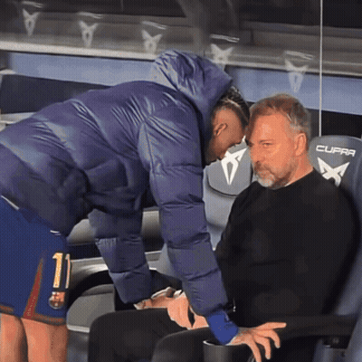 A Man In A Purple Jacket Is Talking To Another Man In A Black Shirt Who Is Sitting In A Cupra Chair GIF