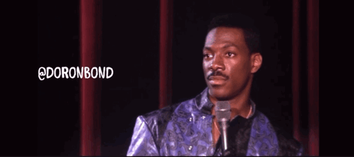 A Man In A Purple Jacket Stands In Front Of A Microphone With The Words @Doronbond Behind Him GIF