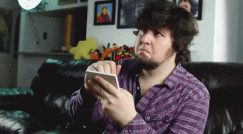 A Man In A Purple Plaid Shirt Is Sitting On A Couch Holding A Cell Phone GIF