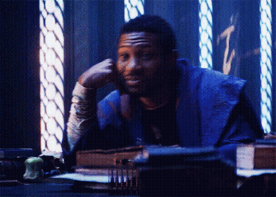 A Man In A Purple Robe Is Sitting At A Desk With His Hand On His Chin GIF
