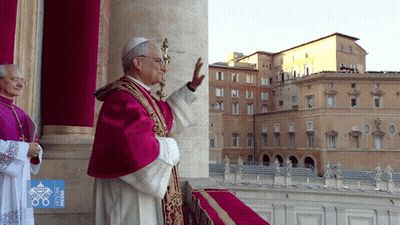 A Man In A Purple Robe Is Standing In Front Of A Building With The Word Vatican On It GIF