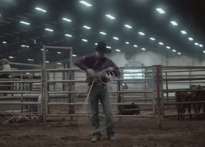 A Man In A Purple Shirt And Hat Is Holding A Lasso In A Rodeo Arena GIF