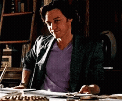A Man In A Purple Shirt Is Sitting At A Desk With A Bookshelf In The Background GIF