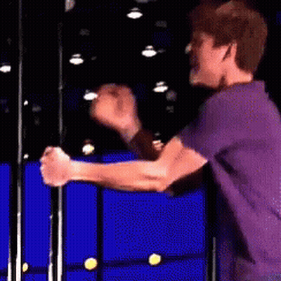 A Man In A Purple Shirt Is Standing In Front Of A Blue Wall With His Hands In The Air . GIF