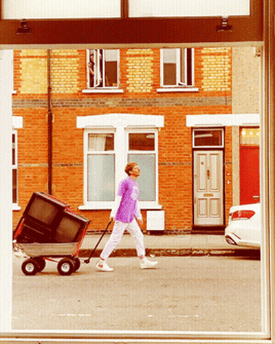 A Man In A Purple Shirt Pushes A Wagon With A Television On It GIF