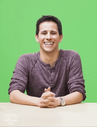 A Man In A Purple Shirt Sits At A Table With His Hands Folded In Front Of A Green Screen GIF