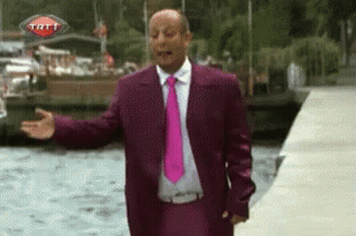 A Man In A Purple Suit And Pink Tie Is Standing In Front Of A Trt Sign GIF