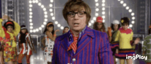 A Man In A Purple Suit And Red Tie Is Standing In Front Of A Group Of Women Dancing . GIF