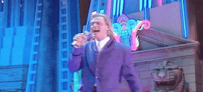 A Man In A Purple Suit Is Singing Into A Microphone On A Stage GIF