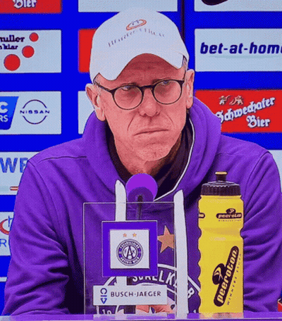 A Man In A Purple Sweatshirt Stands In Front Of A Microphone Next To A Yellow Water Bottle That Says Neeraton GIF