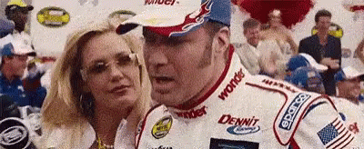 A Man In A Race Car Is Talking To A Woman In Front Of A Crowd Of People . GIF