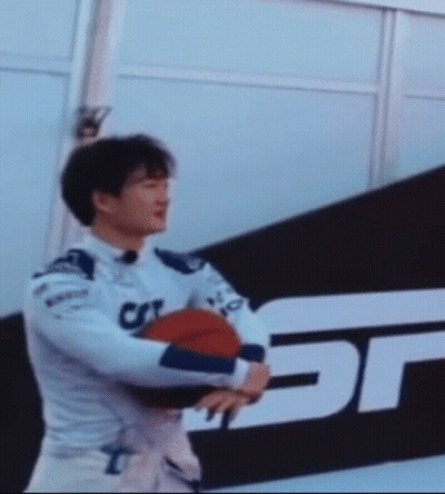 A Man In A Racing Suit Is Holding A Basketball In Front Of A Sign That Has The Letter T On It GIF